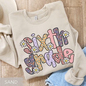 May include: A sand-colored sweatshirt with the words "Sixth Grade" in a colorful, floral design. The letters are filled with various patterns, including flowers and polka dots, and are outlined in a darker color. White daisy accents are placed around the letters.