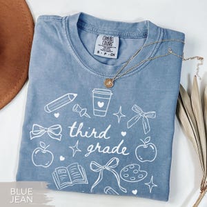 May include: Blue jean-colored t-shirt with white graphic illustrations of school supplies and the words "third grade." A gold necklace with a pendant is draped across the shirt. The shirt is folded and displayed on a white surface.