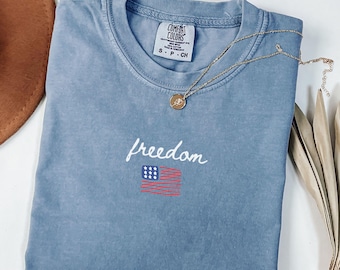 May include: A blue Comfort Colors t-shirt with the word "freedom" in cursive and a small American flag graphic. A gold necklace with a pendant is draped across the shirt. The shirt is folded neatly.