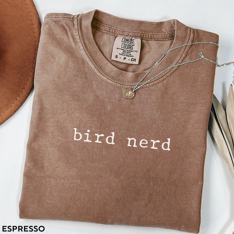 May include: A brown t-shirt with the text "bird nerd" in white lettering. The shirt is made of a soft, comfortable material and has a distressed look. The shirt is shown folded on a white surface.
