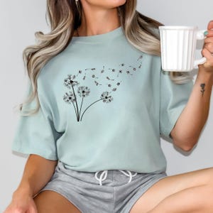 May include: A light blue t-shirt with a black dandelion graphic. The dandelion is blowing in the wind and the seeds are flying away.