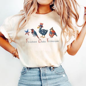 May include: Cream-colored t-shirt featuring three patriotic birds: a flicker, a dodo, and a titmouse. The birds are decorated with stars and stripes, and the text "Flicker Dodo Titmouse" is printed below them.