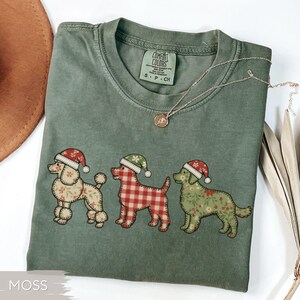 May include: Moss green Comfort Colors t-shirt featuring three dog illustrations wearing Santa hats. The dogs have different patterns: floral, plaid, and solid. A delicate silver necklace with a pendant is draped across the shirt.