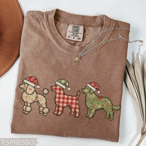 May include: Espresso-colored t-shirt featuring three dogs wearing Santa hats. The dogs have different patterns: floral, plaid, and floral. A silver necklace with a pendant is draped across the shirt. The shirt has a Comfort Colors tag.