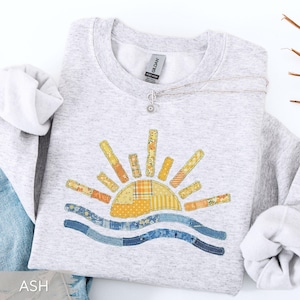 May include: Ash gray sweatshirt with a colorful sun and wave design. The sun is made of patchwork fabric in shades of yellow, orange, and blue. A silver necklace with a small pendant is draped on the sweatshirt.
