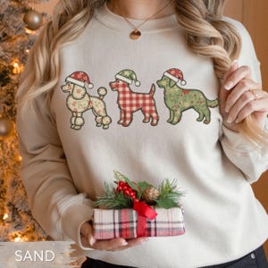 May include: Beige sweatshirt featuring three illustrated dogs wearing Santa hats. The dogs have different patterns: floral, red and white plaid, and green floral. The word "SAND" is printed at the bottom of the image.