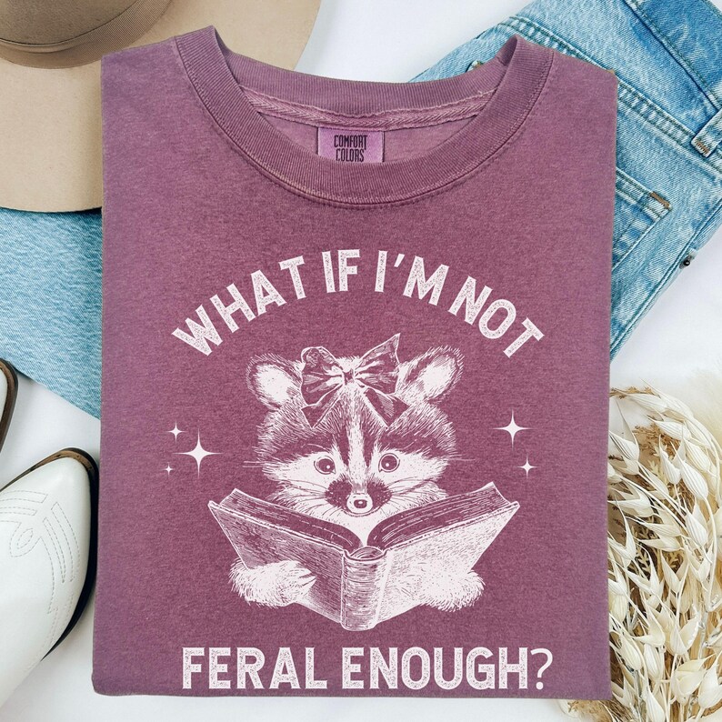 May include: A mauve colored t-shirt with a white graphic of a raccoon wearing a bow and reading a book. The text "What if I'm not feral enough?" is printed on the shirt.