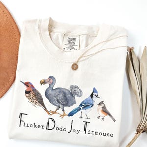 May include: Cream-colored Comfort Colors t-shirt featuring watercolor illustrations of four birds: a flicker, a dodo, a jay, and a titmouse. The names of the birds are printed below them. A gold necklace with a pendant is draped on the shirt.