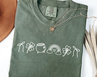 May include: A moss green t-shirt with a white graphic of St. Patrick's Day symbols, including a shamrock, pot of gold, and rainbow. A gold necklace with a pendant rests on the shirt. The shirt is made of Comfort Colors material.