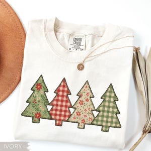 May include: Ivory-colored t-shirt with four Christmas tree appliqués. The trees feature floral and plaid patterns in red, green, and cream colors. A gold necklace with a pendant rests on the shirt. The shirt is labeled "Comfort Colors".