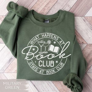 May include: Military green sweatshirt with a white graphic that reads "What Happens at Book Club Stays at Book Club." The design includes martini glasses and an open book. The sweatshirt has a crew neck and long sleeves.