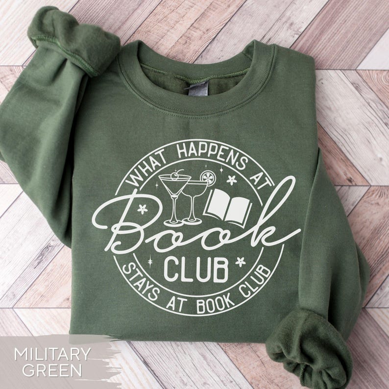 May include: Military green sweatshirt with a white graphic that reads "What Happens at Book Club Stays at Book Club." The design includes martini glasses and an open book. The sweatshirt has a crew neck and long sleeves.