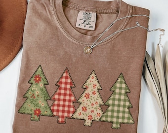 Könnte beinhalten: Espressofarbenes T-Shirt mit vier Weihnachtsbaum-Applikationen. Die Bäume haben Blumen- und Karomuster in Rot, Grün und Creme. Eine silberne Halskette mit Anhänger liegt auf dem Shirt. Das Shirt hat ein Comfort Colors-Etikett.