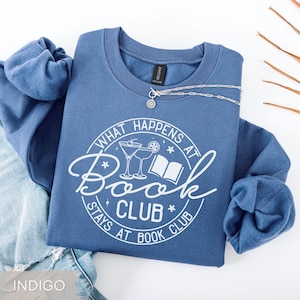 May include: Indigo blue sweatshirt with the text "WHAT HAPPENS AT BOOK CLUB STAYS AT BOOK CLUB" in a white circular design. The design includes cocktail glasses and a book. A silver necklace with a sunburst pendant is draped across the sweatshirt.