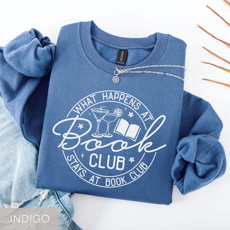 May include: Indigo blue sweatshirt with the text "WHAT HAPPENS AT BOOK CLUB STAYS AT BOOK CLUB" in a white circular design. The design includes cocktail glasses and a book. A silver necklace with a sunburst pendant is draped across the sweatshirt.