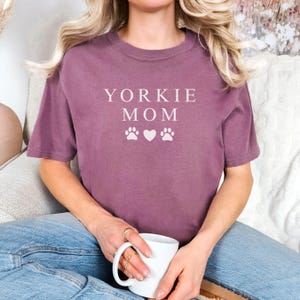 May include: A mauve colored t-shirt with the text "Yorkie Mom" and two paw prints and a heart in white.