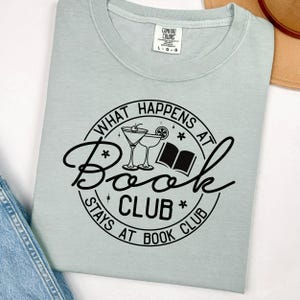 May include: A light blue t-shirt with a black circle graphic that says "What Happens at Book Club Stays at Book Club". The graphic also includes a book, two martini glasses, and stars.