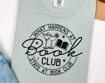 May include: A light blue t-shirt with a black circle graphic that says "What Happens at Book Club Stays at Book Club". The graphic also includes a book, two martini glasses, and stars.