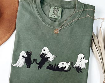 May include: Moss green Comfort Colors t-shirt featuring a playful design of black cats interacting with white ghosts. A delicate gold necklace with a pendant rests on the shirt. The shirt is a size S-P-CH.