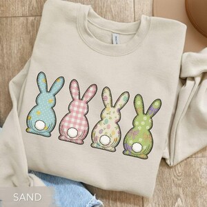 May include: Sand-colored sweatshirt featuring four Easter bunny designs. Each bunny has a unique pattern: blue polka dots, pink gingham, floral with Easter eggs, and green floral. The bunnies have white fluffy tails and black outlines.