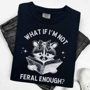 May include: Black t-shirt with a white graphic of a raccoon wearing a bow and reading a book. The text "What if I'm not feral enough?" is printed on the shirt.