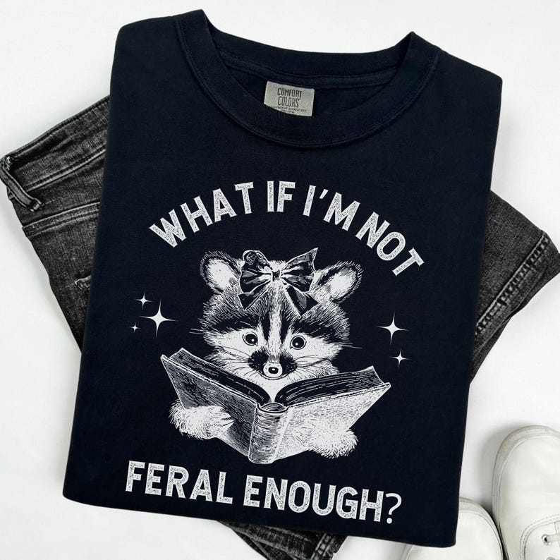 May include: Black t-shirt with a white graphic of a raccoon wearing a bow and reading a book. The text "What if I'm not feral enough?" is printed on the shirt.