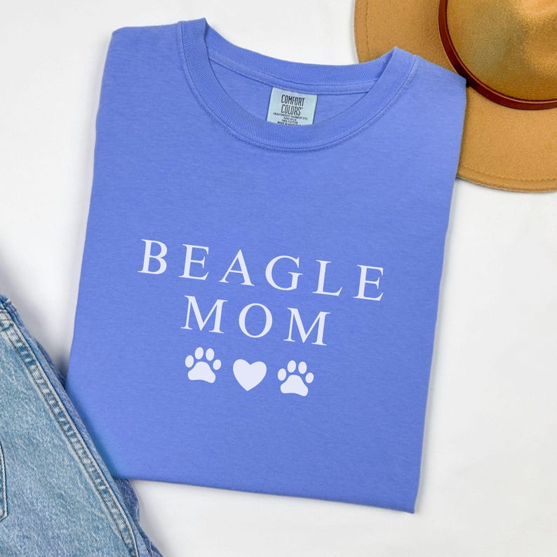 K&ouml;nnte beinhalten: Ein hellblaues T-Shirt mit dem Text "BEAGLE MOM" in wei&szlig;en Buchstaben. Zwei wei&szlig;e Pfotenabdr&uuml;cke und ein wei&szlig;es Herz befinden sich unter dem Text.