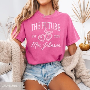 May include: A bright pink t-shirt with the text "THE FUTURE EST 2026 Mrs. Johnson" in white script. The design includes two heart-shaped graphics with the letters "T" and "S". The shirt is paired with denim shorts.