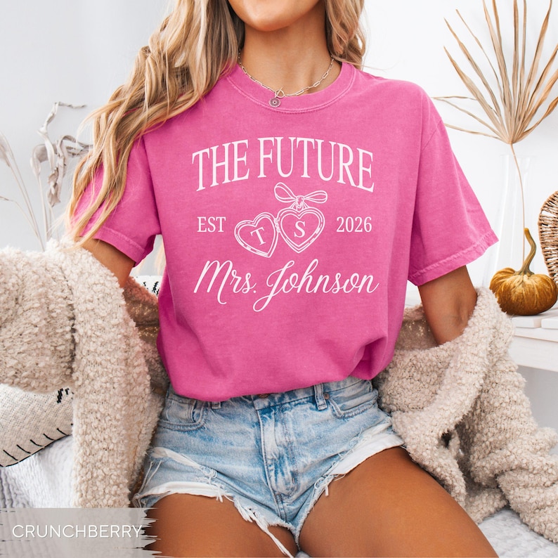 May include: A bright pink t-shirt with the text "THE FUTURE EST 2026 Mrs. Johnson" in white script. The design includes two heart-shaped graphics with the letters "T" and "S". The shirt is paired with denim shorts.