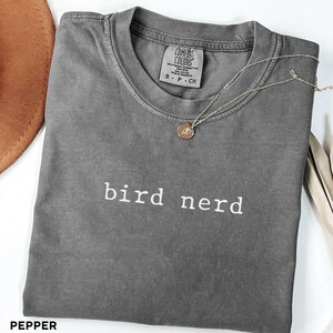 May include: A grey t-shirt with the text "bird nerd" printed in white on the front. The shirt is made of a soft, comfortable material and has a relaxed fit.