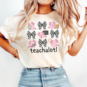 May include: Cream-coloured t-shirt with a design of pink axolotls, black and white checkered bows, books, a globe, a laptop, a clock, and a chalkboard with "ABC". The word "teachalotl" is printed below the axolotls.