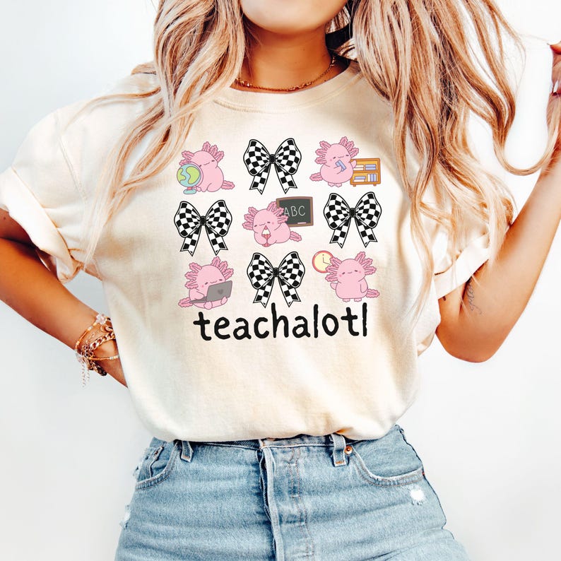 May include: Cream-coloured t-shirt with a design of pink axolotls, black and white checkered bows, books, a globe, a laptop, a clock, and a chalkboard with "ABC". The word "teachalotl" is printed below the axolotls.