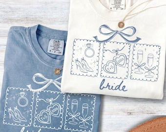 May include: Two Comfort Colors t-shirts, one blue and one cream, with wedding-themed graphics. The blue shirt says "bridesmaid" and the cream shirt says "bride". Each shirt features a bow and three framed illustrations: a ring, shoes, hearts, and champagne flutes.