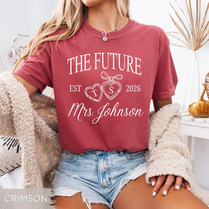 May include: Crimson-colored t-shirt with the text "THE FUTURE EST 2026 Mrs. Johnson" in white script. The design includes two heart-shaped pendants with the letters "T" and "S". The shirt is short-sleeved.