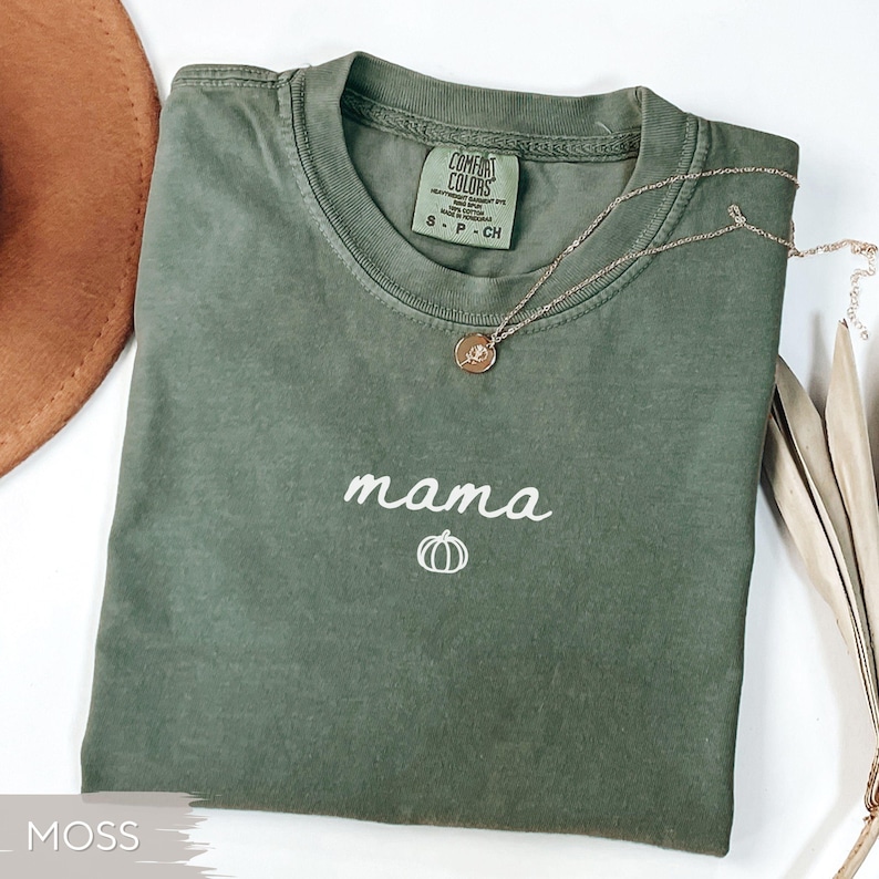 May include: Moss green t-shirt with the word "mama" in white script and a pumpkin graphic. A gold necklace with a pendant rests on the shirt. The shirt is folded and has a tag that says "Comfort Colors".