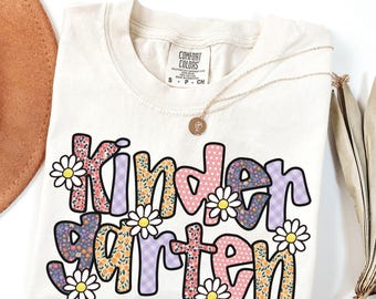 May include: Ivory t-shirt featuring the word "Kindergarten" in a colourful floral and polka dot design. The letters are outlined in black and decorated with daisy accents. A gold necklace with a pendant is draped on the shirt.