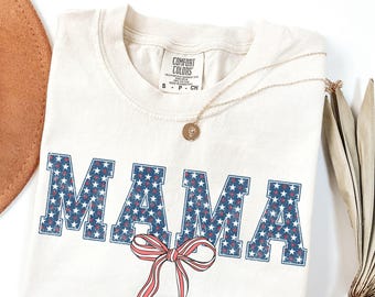 Puede incluir: Una camiseta de color crema con un gráfico de "MAMA" azul y rojo desgastado con estrellas blancas. El gráfico tiene un lazo rojo y blanco debajo.