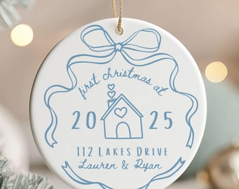 May include: A white ceramic Christmas ornament with a blue ribbon design and the text "first christmas at 2025". The ornament features a house graphic with a heart and the address "112 LAKES DRIVE Lauren & Ryan".