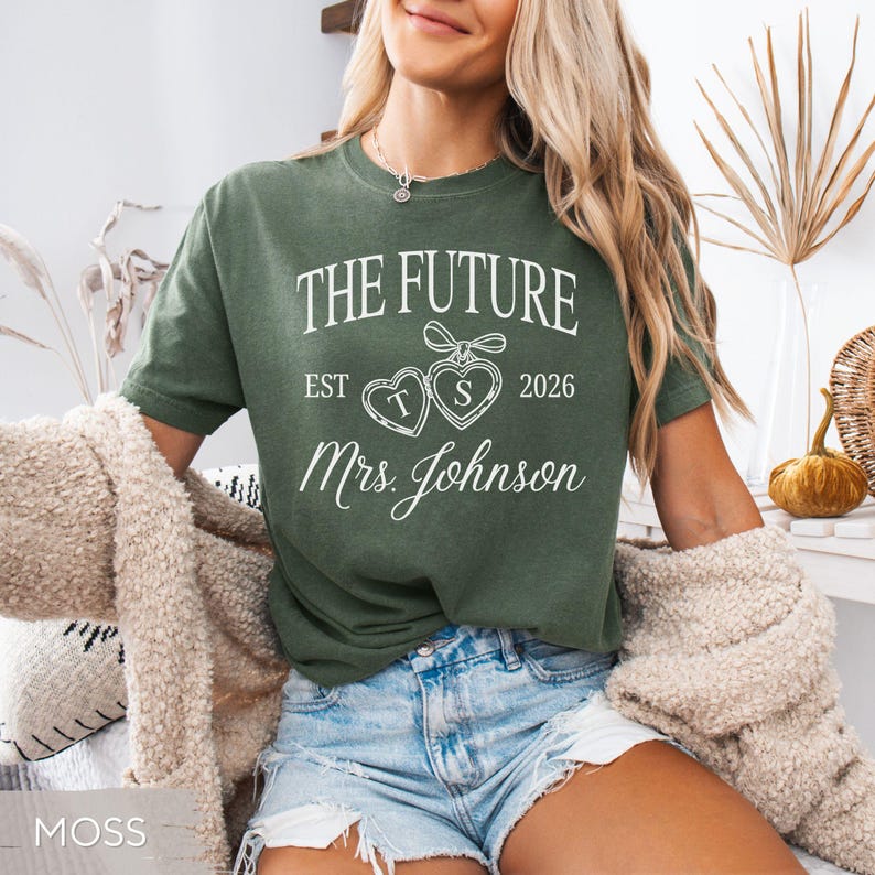 May include: Moss green t-shirt with the text "THE FUTURE EST 2026 Mrs. Johnson" in white. The design includes two heart-shaped lockets with the letters "T" and "S". The shirt is a casual style.