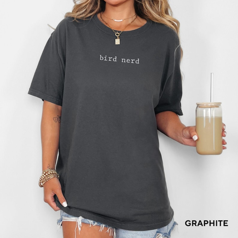 May include: An oversized t-shirt in a dark grey colour with the text "bird nerd" printed in white on the front. The shirt is being modelled by a person wearing denim shorts.