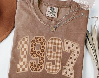 May include: A brown short-sleeve t-shirt featuring the number 1997 in large block letters. Each digit has a different pattern: a checkerboard, leopard print, gingham plaid, and polka dots. A silver necklace with a round pendant rests on the shirt.