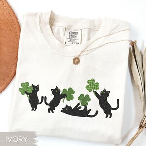 May include: Ivory-colored Comfort Colors t-shirt featuring four black cat illustrations holding green shamrocks. A gold necklace with a pendant rests on the shirt. The shirt has a crew neck and short sleeves.