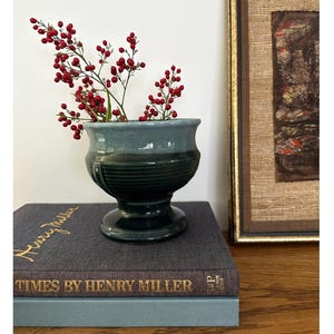 May include: A two-toned teal ceramic pedestal vase with a flared rim and horizontal ribbed detailing on the body. The vase contains a sprig of red berries. It is positioned on a dark gray book titled Times by Henry Miller.