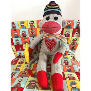 May include: A gray sock monkey doll with a red mouth, red hands, and red feet. It wears a multicolored striped knit cap and has a red heart patch on its chest. A yellow banana is held in one hand, and thin colorful ribbons are tied around its neck.