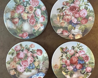 Pode incluir: Um conjunto de quatro pratos redondos de cerâmica, cada um com uma ilustração detalhada de um buquê de rosas cor-de-rosa, vermelhas e amarelas num vaso branco. O fundo de cada prato retrata uma cena de jardim suave e pitoresca.