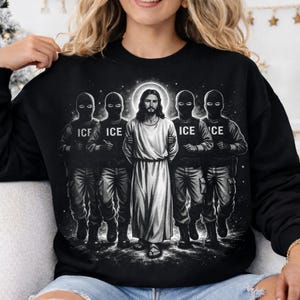 Puede incluir: Sudadera negra con un gráfico de Jesús rodeado por cuatro figuras con equipo táctico, cada una con la palabra "ICE" en sus uniformes. Jesús está representado con una túnica blanca y un halo.