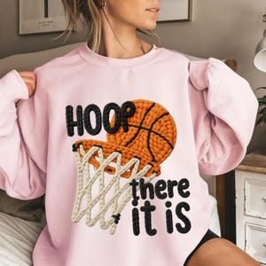 Puede incluir: Una sudadera rosa claro con un diseño de baloncesto. El diseño presenta una pelota de baloncesto, un aro y las palabras "HOOP there it is" en negro. La pelota es naranja con líneas negras, y la red es beige.