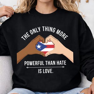 May include: Black sweatshirt with the text "THE ONLY THING MORE POWERFUL THAN HATE IS LOVE." The design features two hands forming a heart around the Puerto Rican flag.