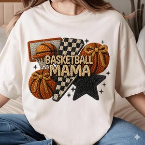 Puede incluir: Camiseta color crema con un diseño temático de baloncesto. El diseño incluye una pelota de baloncesto, una red, un patrón de cuadros y una estrella, con el texto "BASKETBALL MAMA" en una fuente texturizada. La camiseta es de un material suave.