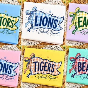 Puede incluir: Seis paneles de tela rectangulares con gráficos de banderines con patrones de cuadros y lazos. Los diseños incluyen el texto GATORS, LIONS, EAGLES, TIGERS y BEASRS, cada uno acompañado por la frase School Name en una fuente cursiva.